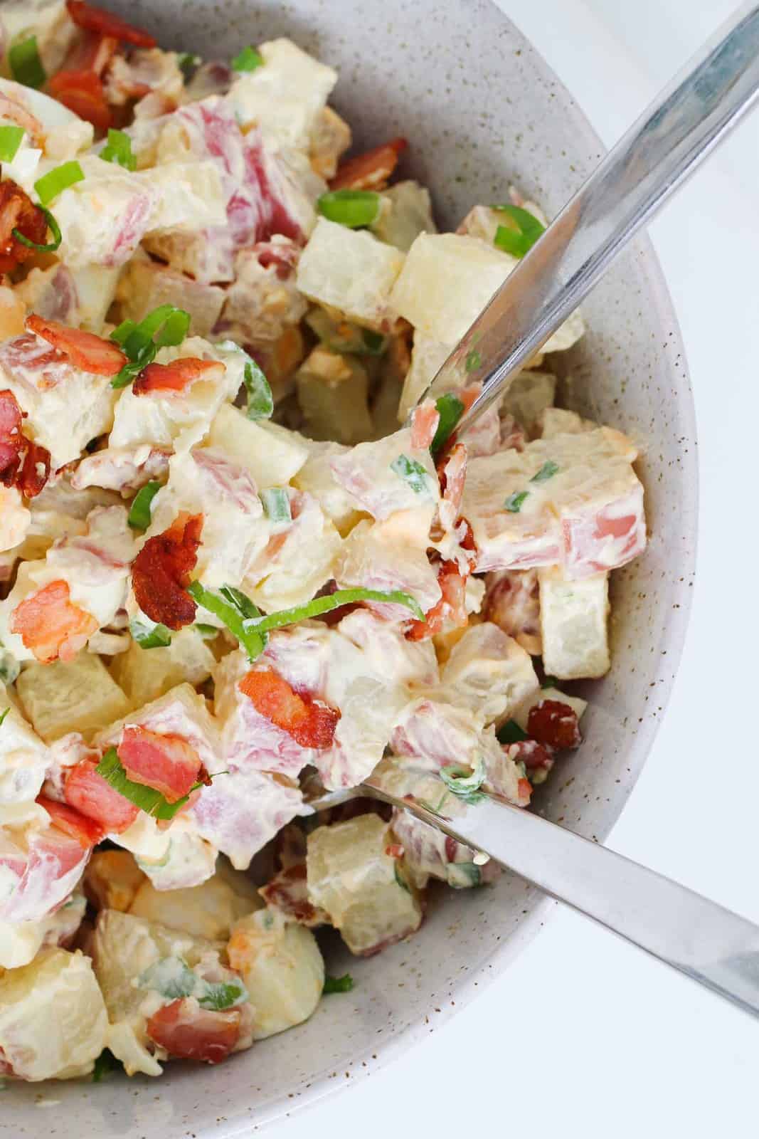 Salad servers in a bowl of potatoes, bacon, eggs, spring onions with a creamy dressing.