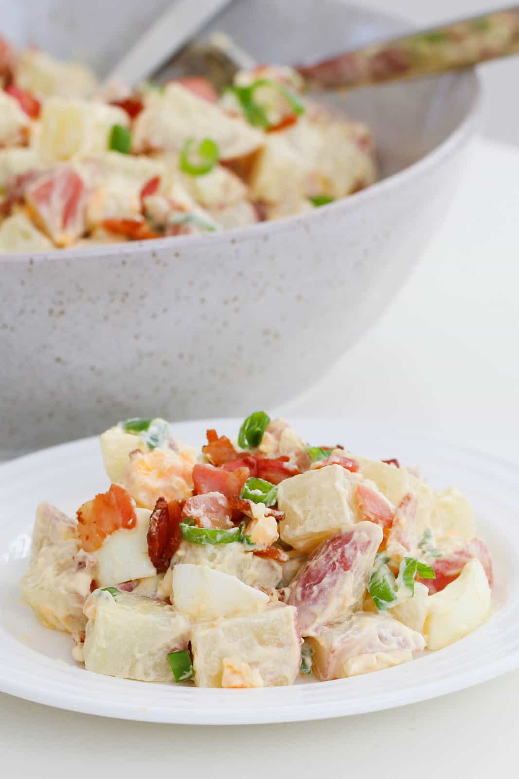 A plate of potato salad with egg, bacon and spring onions, and a bowl of potato salad in the background