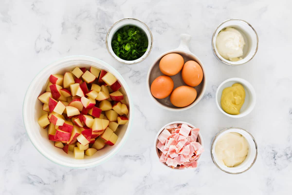 All the ingredients for potato salad in individual bowls on a marble bench