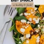 Two silver salad servers next to a bowl of spinach leaves with roasted pumpkin pieces, crumbked feta and pine nuts.