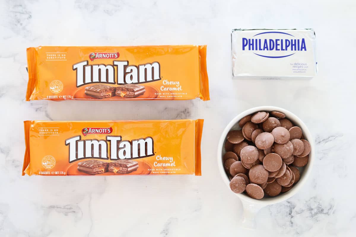 A block of cream cheese, milk chocolate buttons and two packets of caramel Tim Tams on a white marble board.