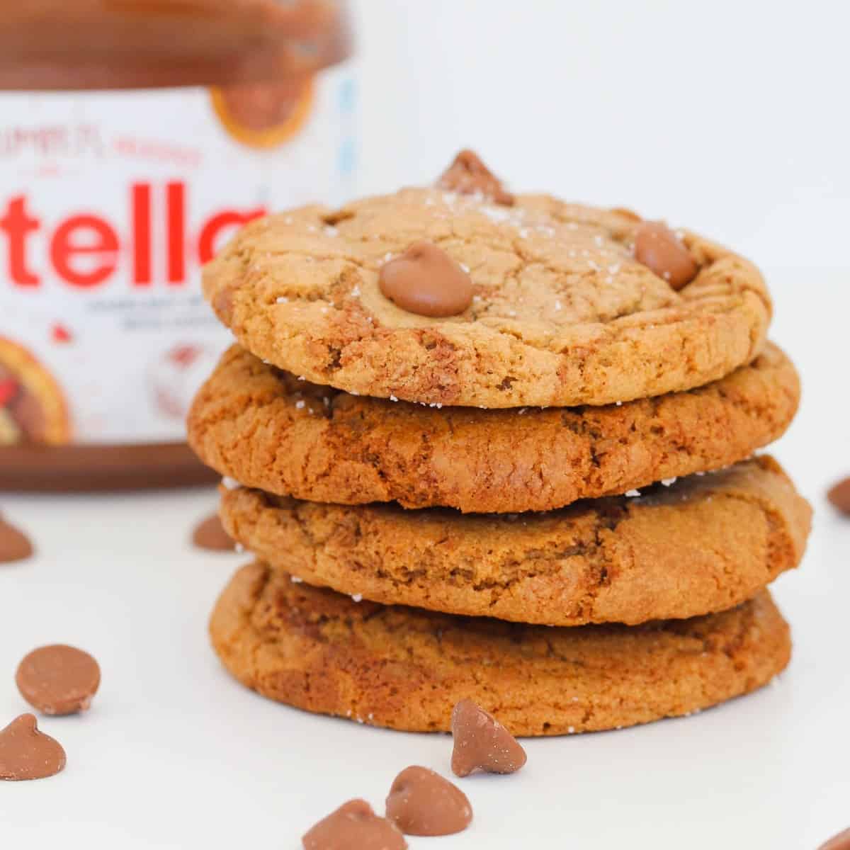 Four brown cookies in a stack with chocolate chips surrounding them and a jar of Nutella.