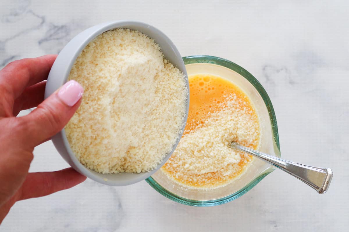 Pouring grated parmesan cheese into beaten egg mixture in glass measuring cup.