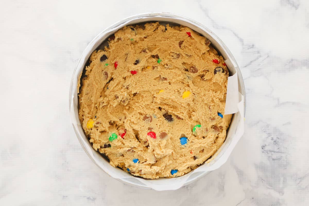 Cookie cake batter with chocolate chips and M&M's in a lined cake tin.