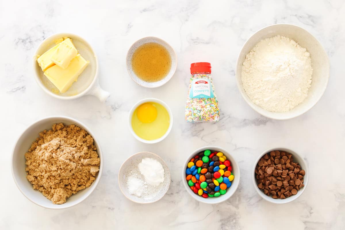 Ingredients for cookie cake with M&M's and chocolate chips in individual bowls on a marble bench top.