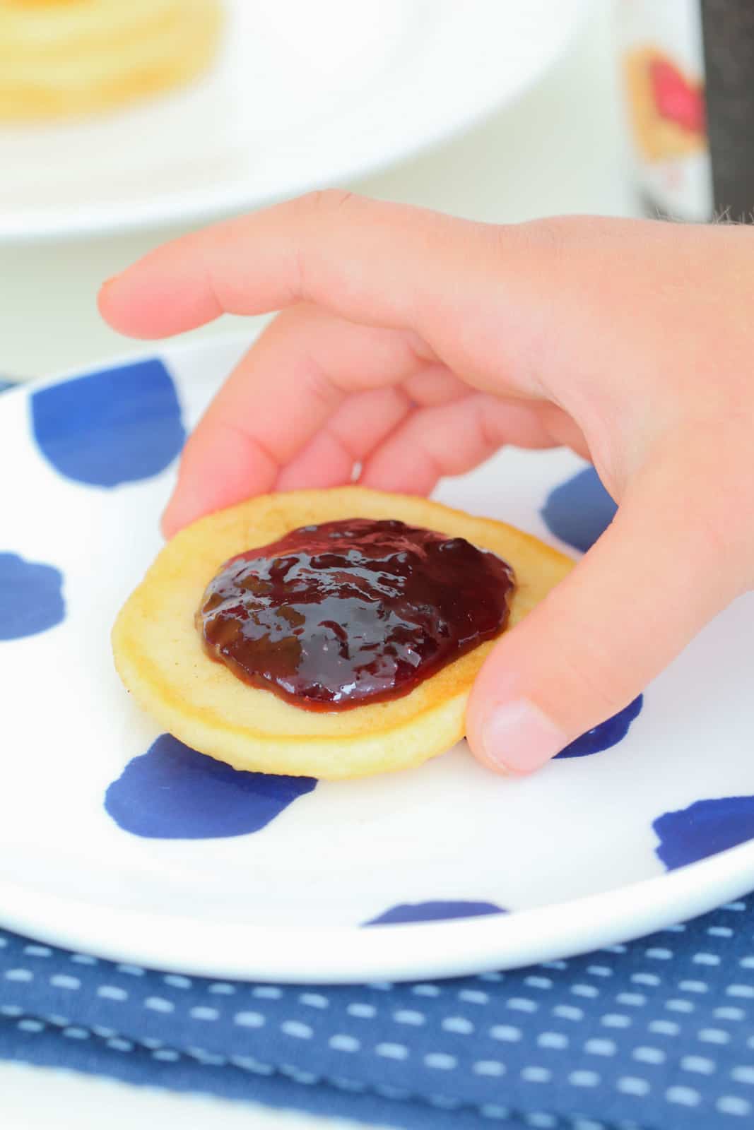 A child's hand holding a pikelet topped with jam.