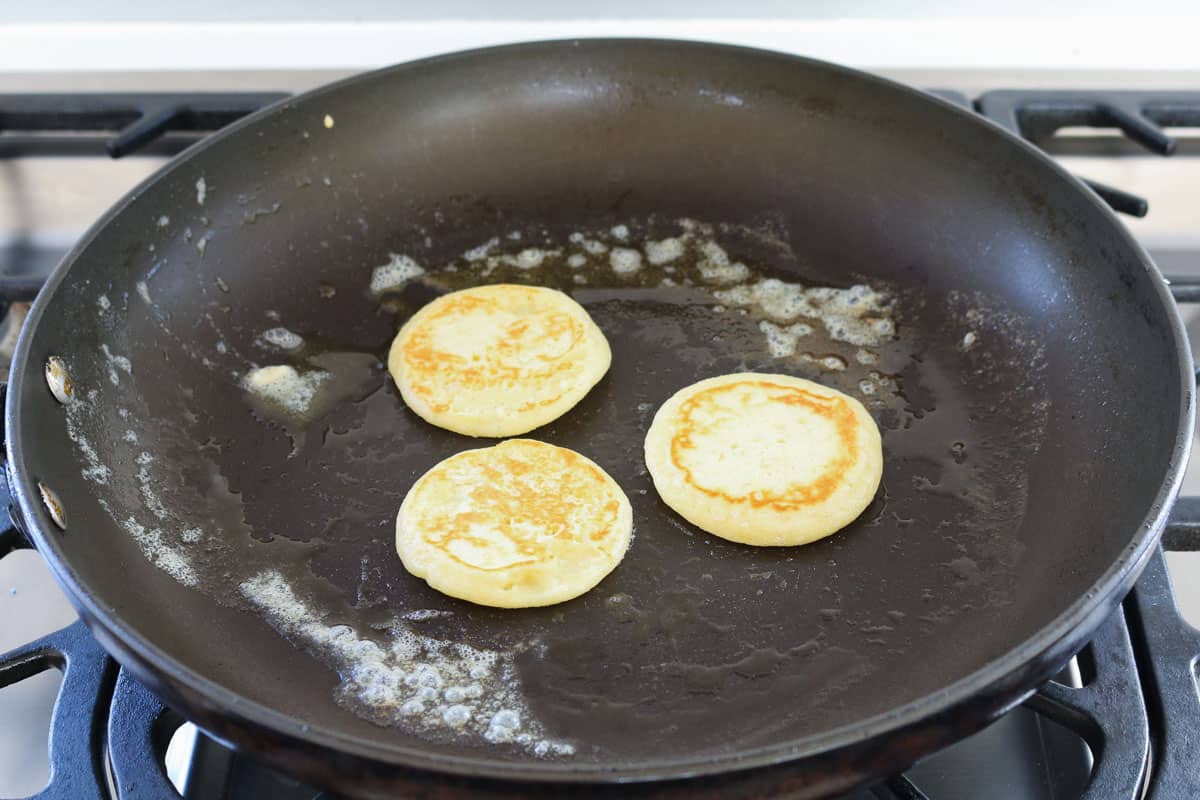 Three golden pikelets cooking in a frying pan.