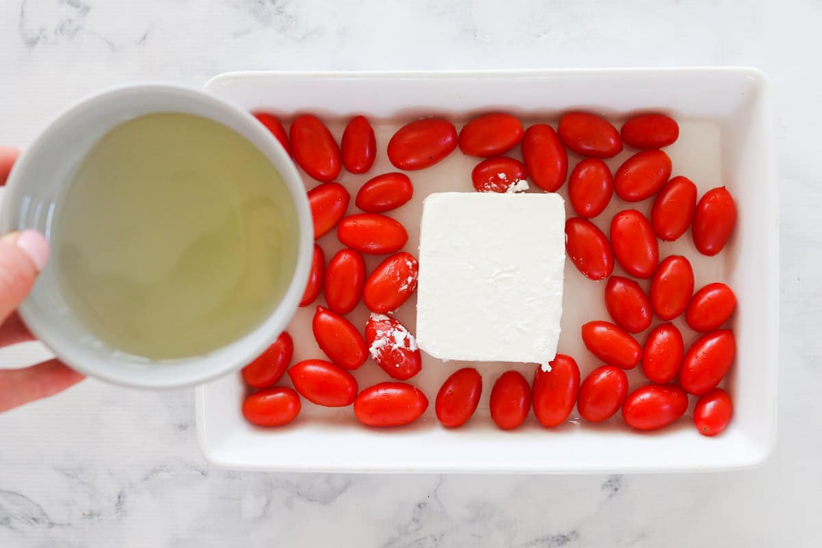 Olive oil being poured onto feta and tomatoes in a baking dish.