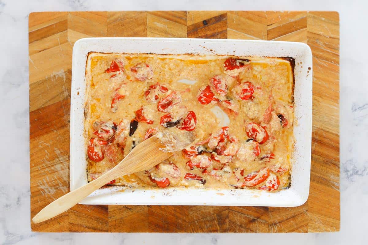 A wooden spoon stirring a baking dish with a creamy tomato and feta sauce.