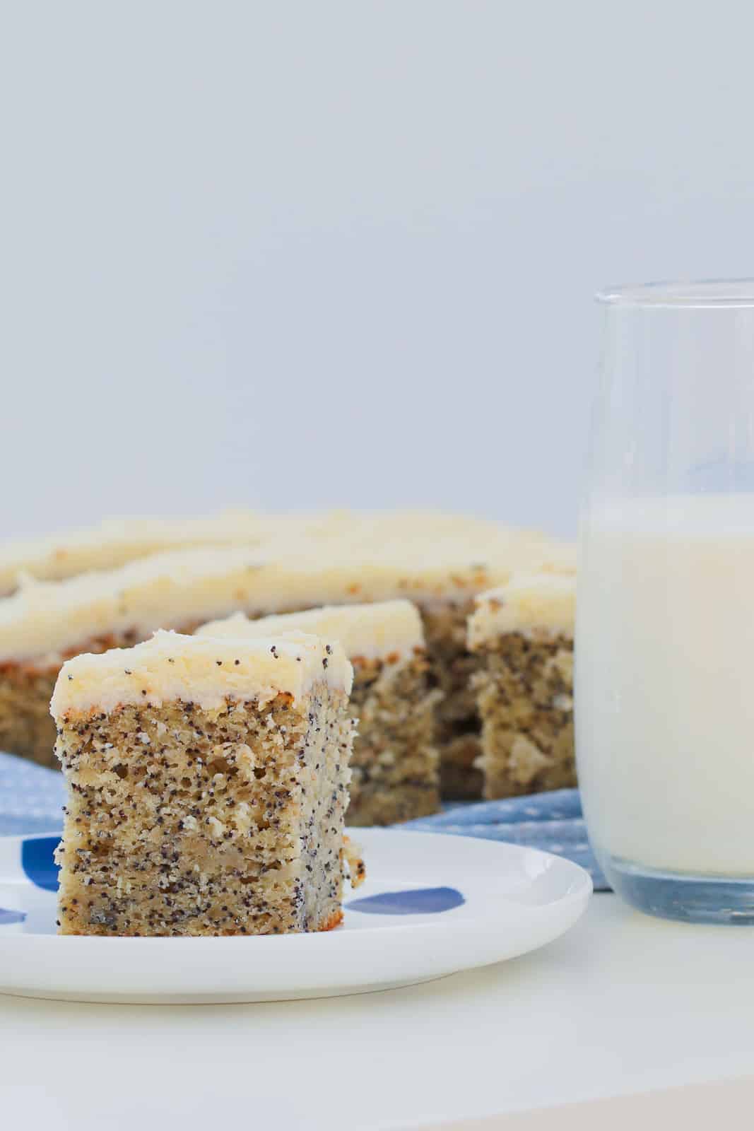 A piece of frosted banana poppyseed cake on a white and blue plate, with the rest of the sliced cake blurred in the background