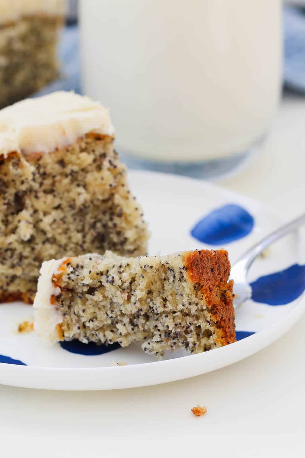 A split piece of frosted poppy seed cake on a white & blue plate, with a glass of milk and more cake on a blue and white towel in the background.