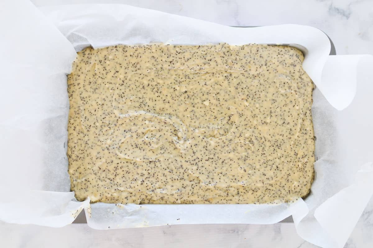 A baking paper lined rectangular baking tin filled with a poppy seed cake batter.