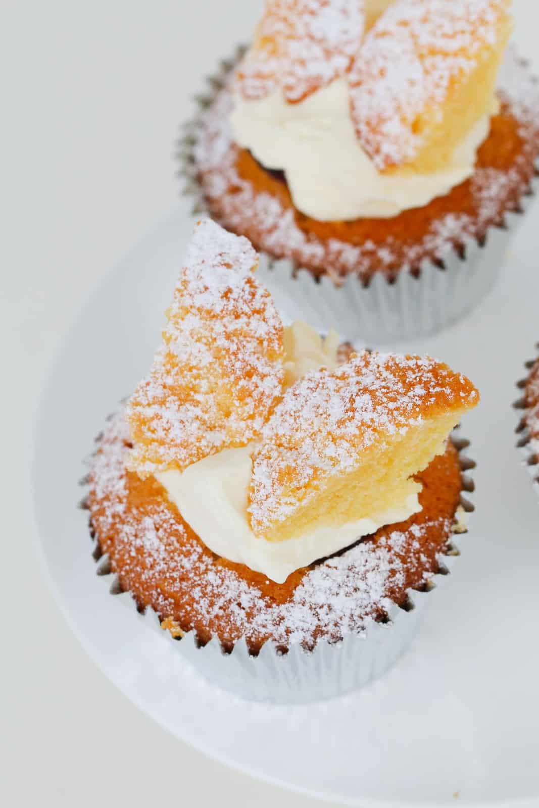 A butterfly cupcake filled with jam and cream and dusted with icing sugar on a white cake stand.