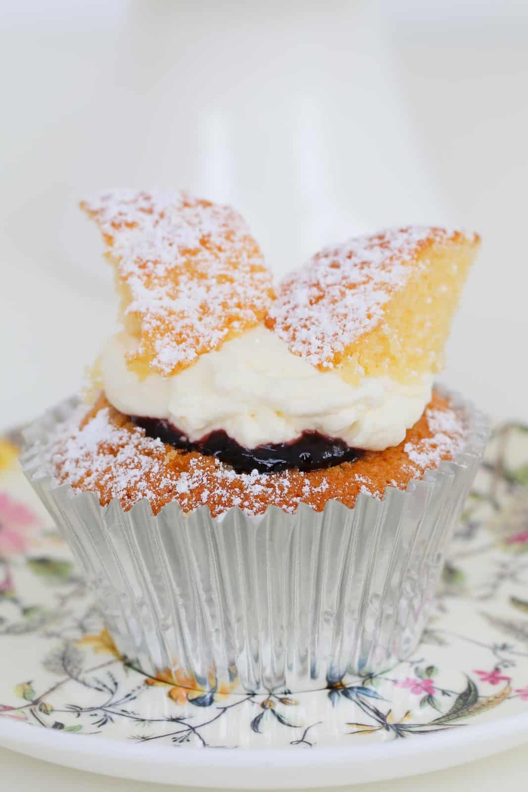 A close up of a silver cupcake case holding a vanilla fairy cupcake with jam and cream, and dusted with icing sugar.