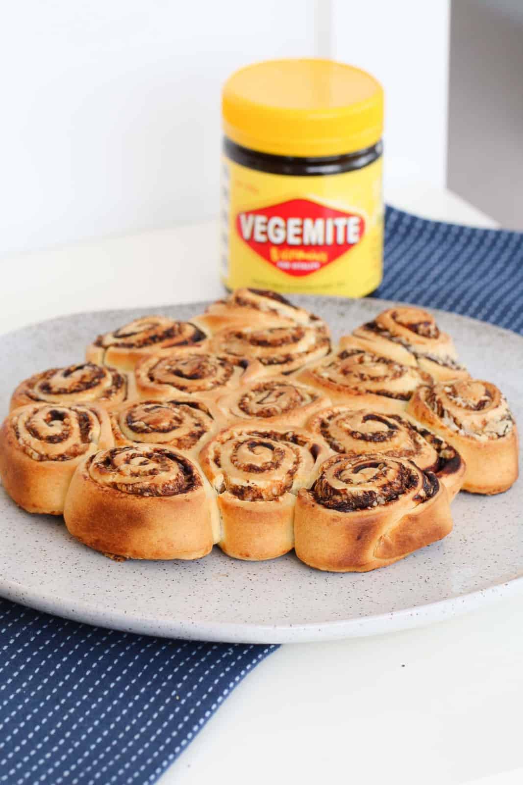 A batch of baked scrolls on a speckled plate on a blue and white tea towel, with a jar of Vegemite in the background.