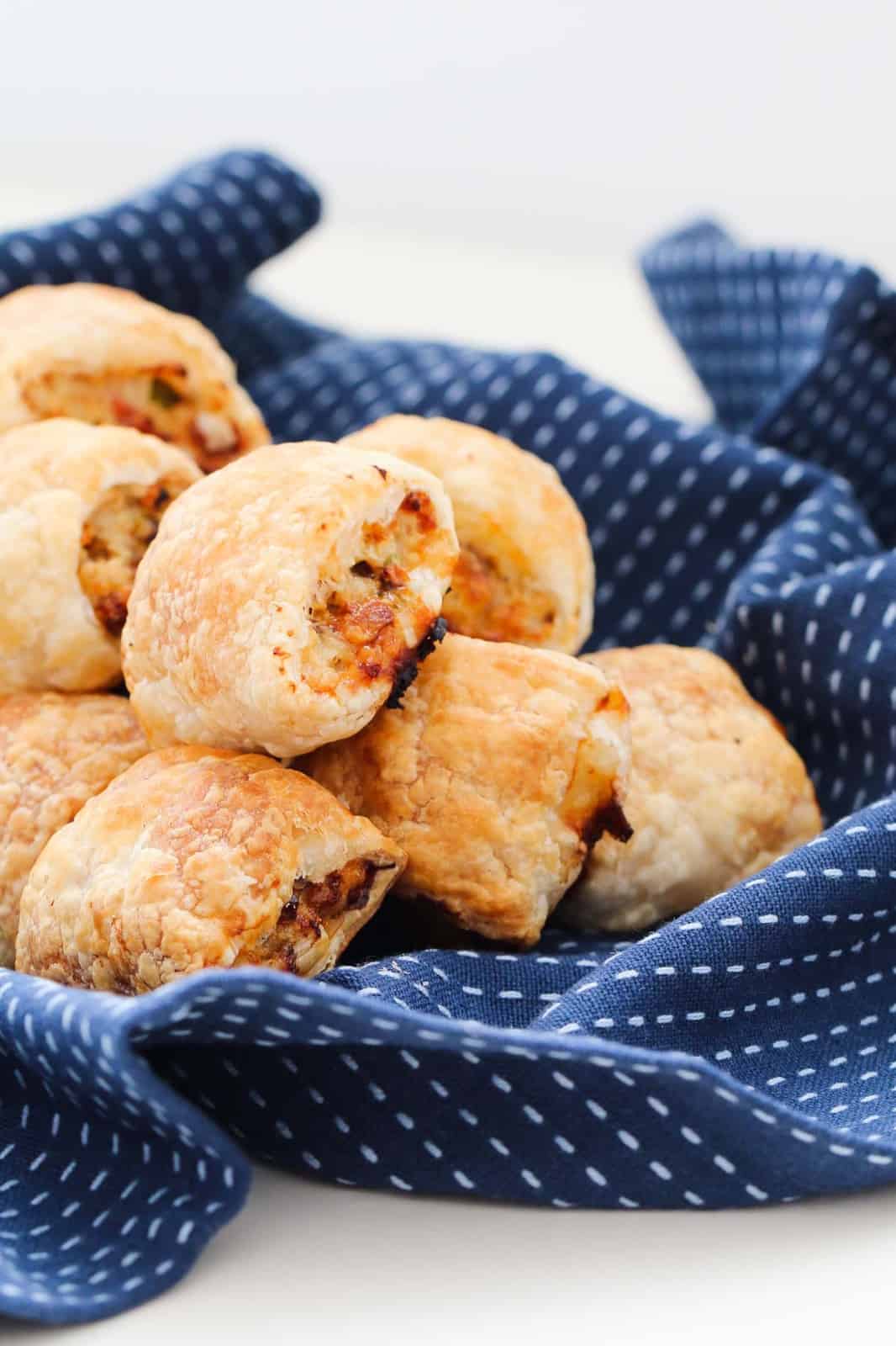 A batch of golden chicken, cheese and bacon sausage rolls piled on a blue tea towel.