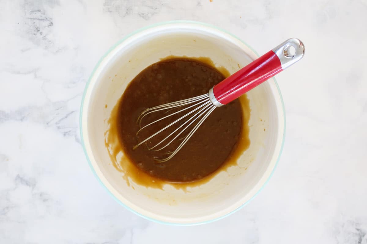 Ingredients, including brown sugar, being mixed with a red handled whisk into a melted butter mixture in a white bowl.