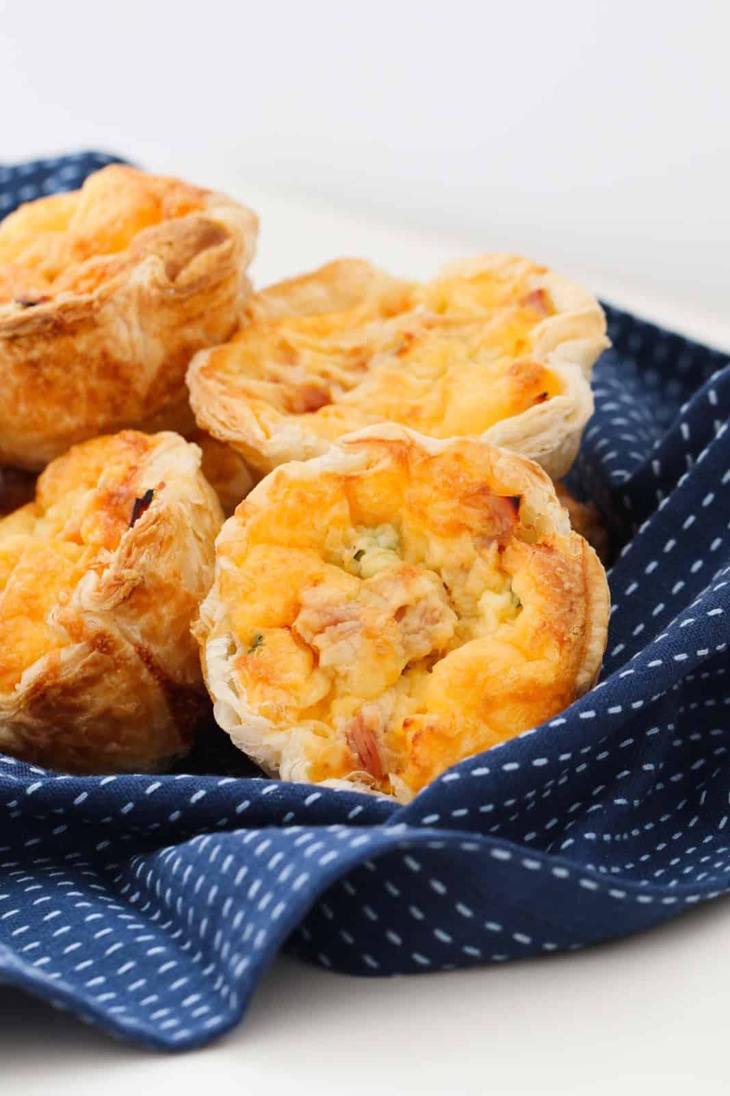 Four mini corn, ham and cheese quiches with a puff pastry crust arranged on a blue patterned tea towel.