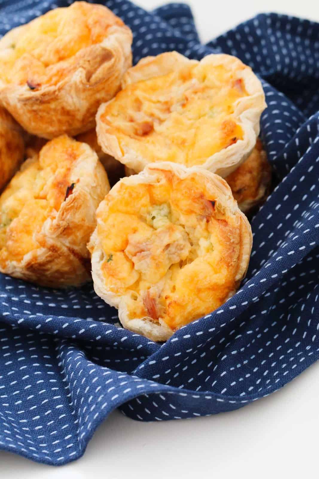 A batch of individual serve quiches with a puff pastry crust arranged on a blue patterned tea towel.