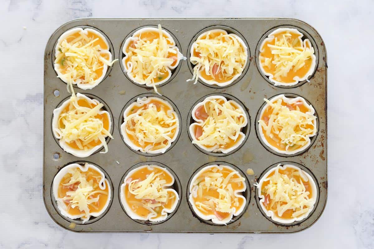 Grated cheese sprinkled over fillings of corn, ham, spring onions and beaten egg in a muffin tray