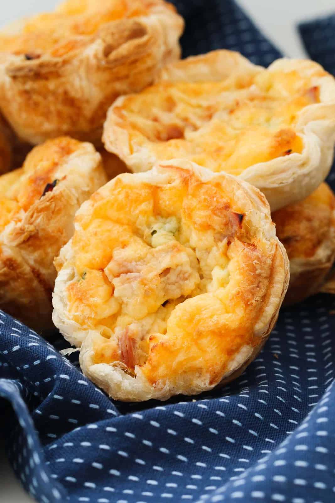 Individual serve golden mini corn, ham and cheese quiches with a puff pastry crust arranged on a blue patterned tea towel.