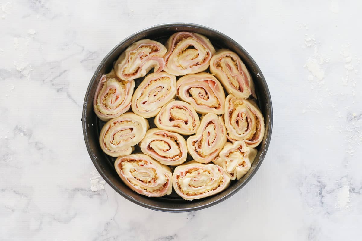 Pinwheels of homemade dough with ham, mayonnaise and cheese inside, placed in a round baking tin. 