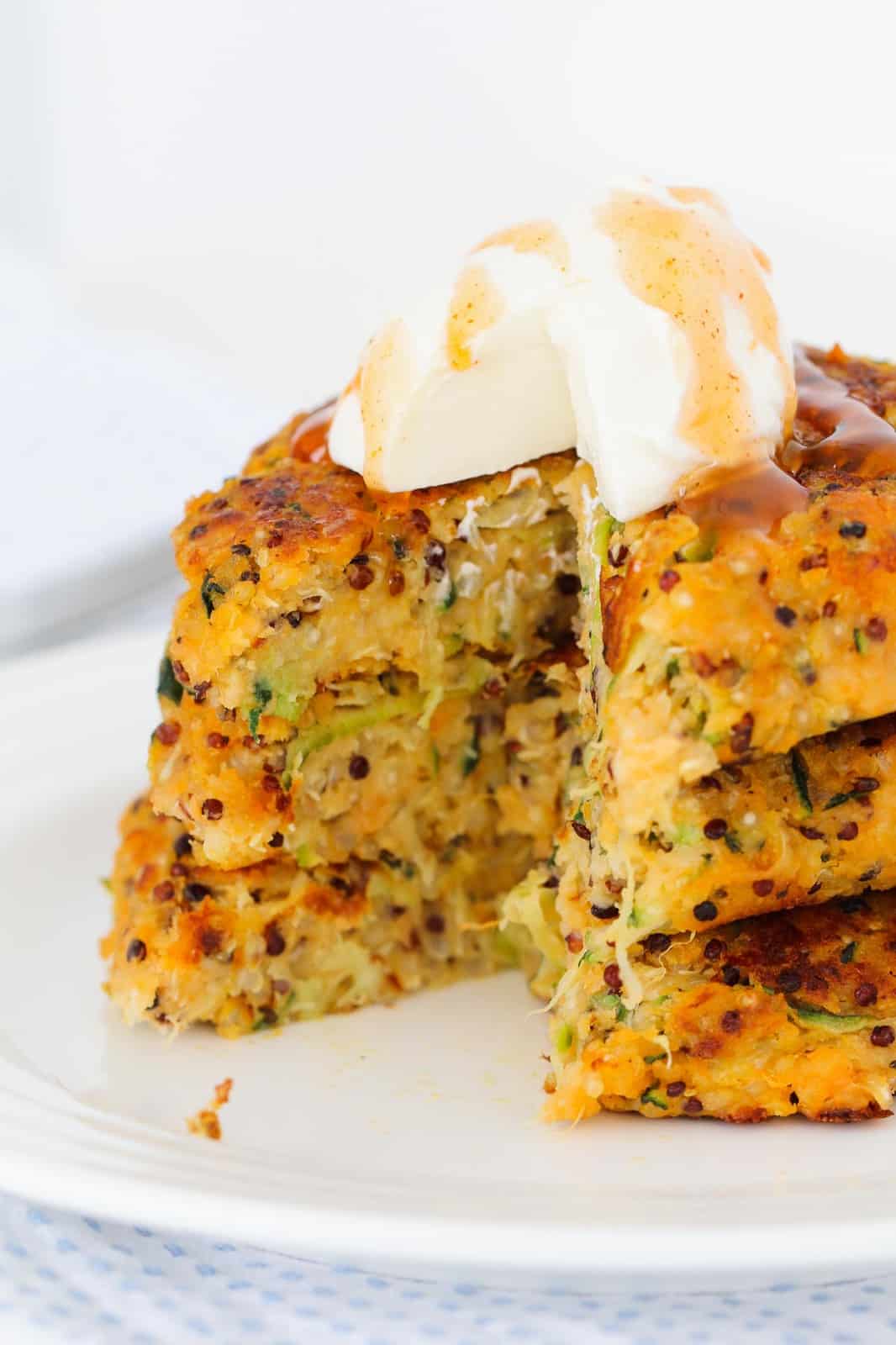 Close up of cheesy quinoa, zucchini, and sweet potato fritters in a stack topped with sour cream and drizzled with sweet chili sauce
