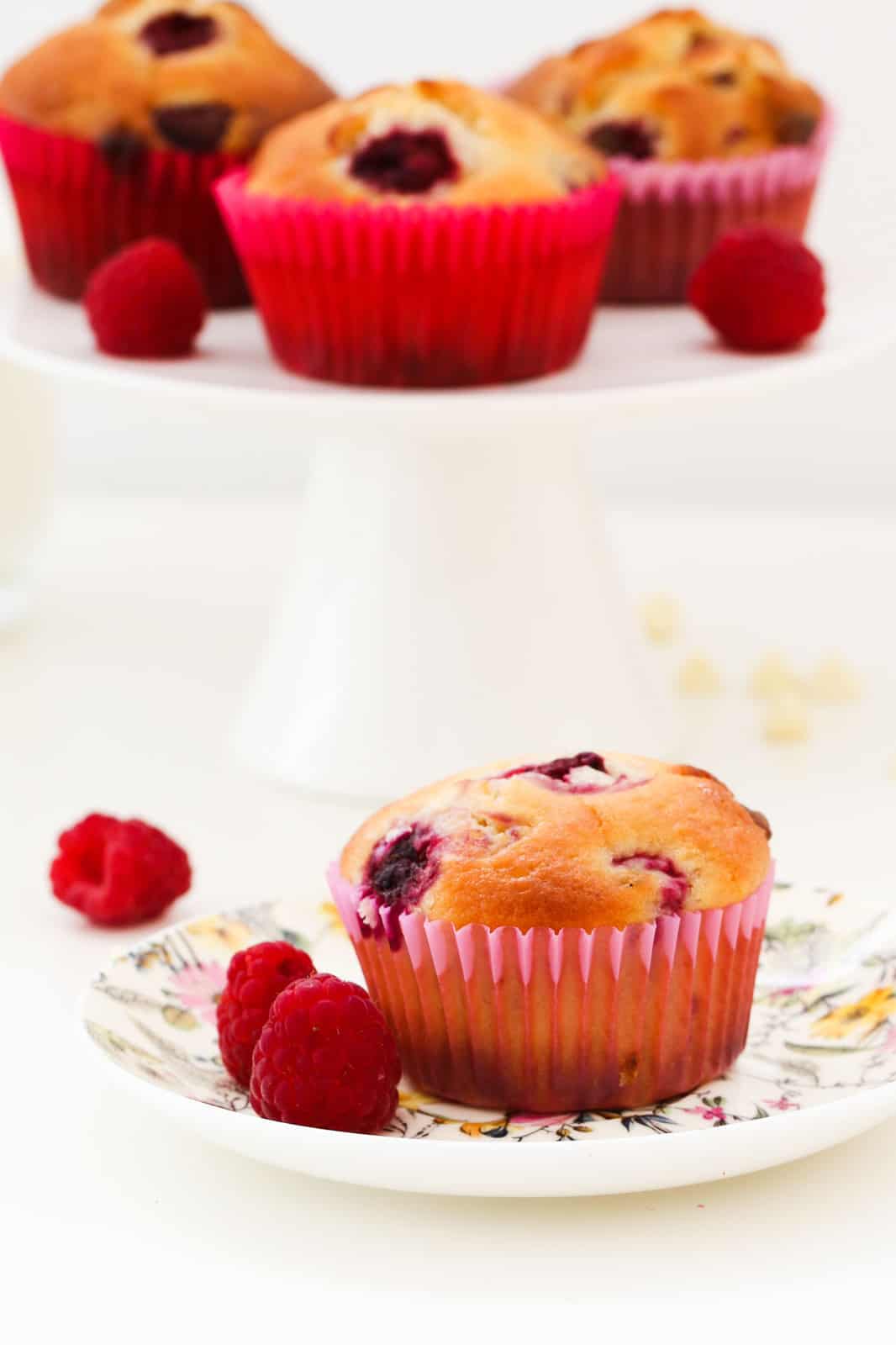A raspberry muffin and some fresh raspberries on a floral plate in the foreground, and a blurred cake stand with three muffins in the background
