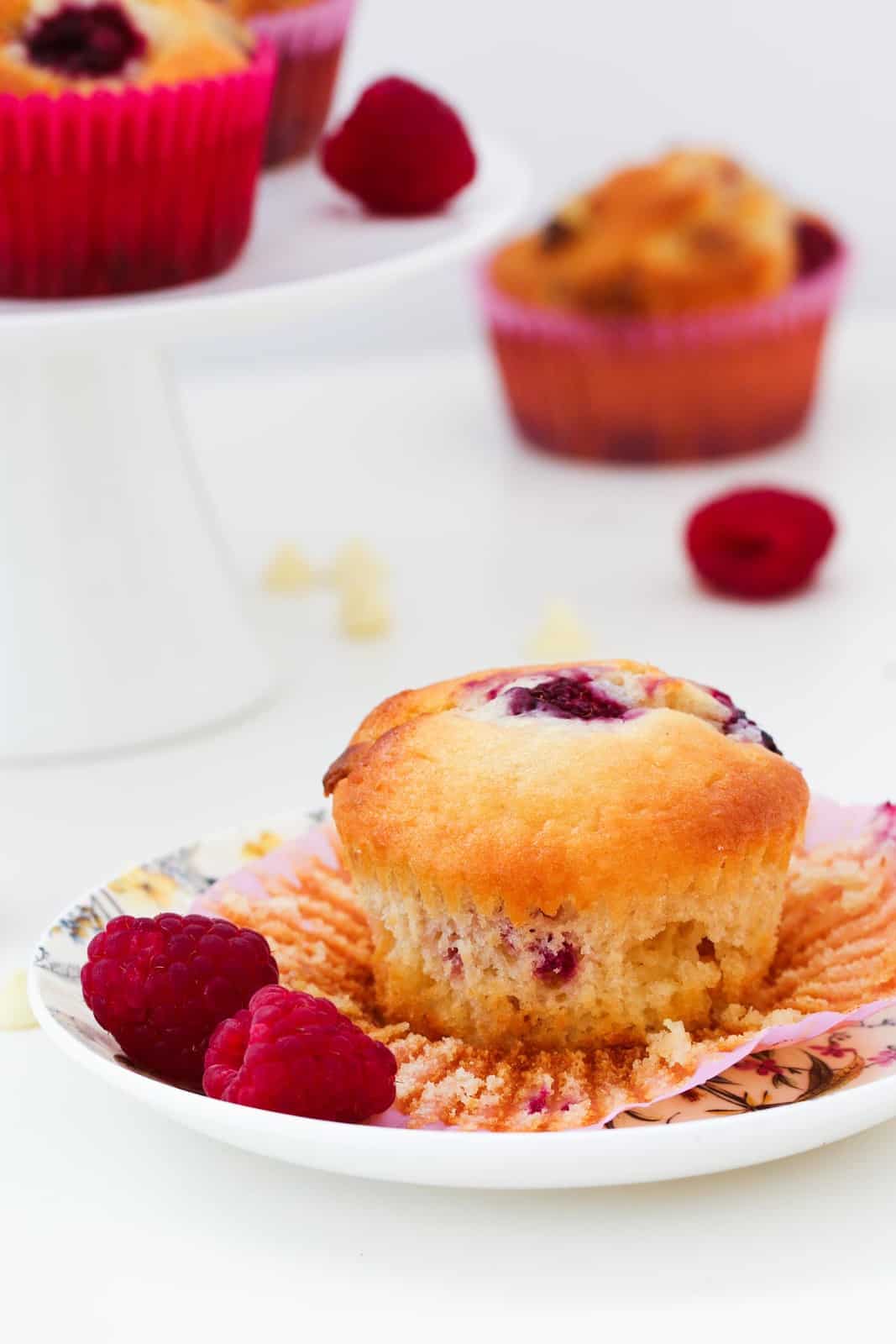 A berry muffin with the paper case opened and served with fresh raspberries on a floral plate.
