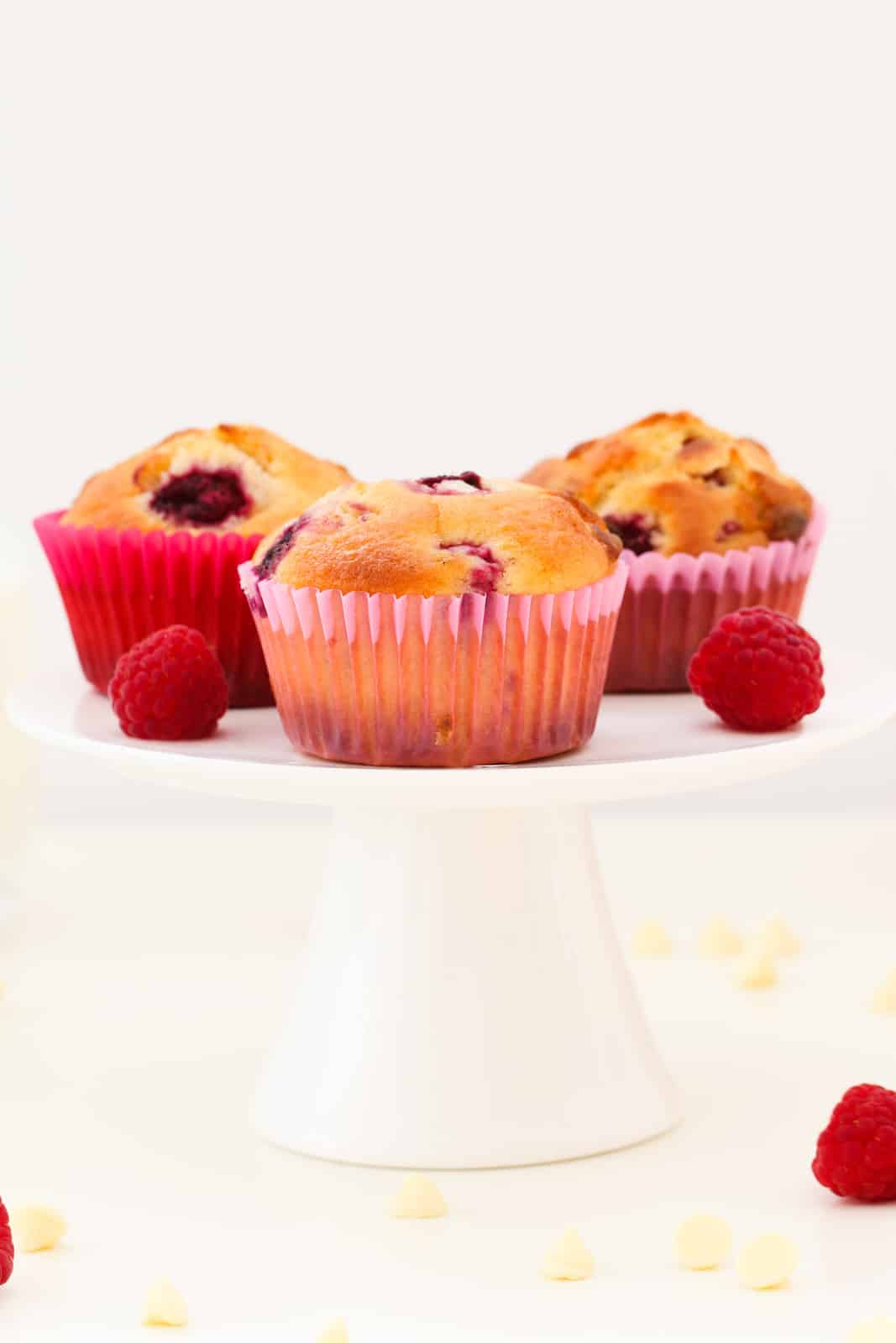 Muffins in pink and red paper cases and two fresh raspberries on a white cake stand.