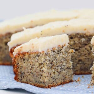 A piece of frosted poppy seed cake on a blue and white tea towel in front of the rest of the sliced cake.