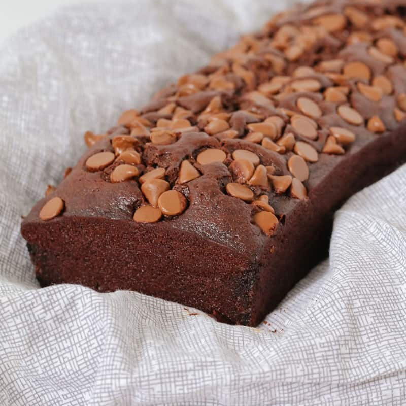 A chocolate loaf with chocolate chips on top, sitting on a white and grey patterned tea towel.