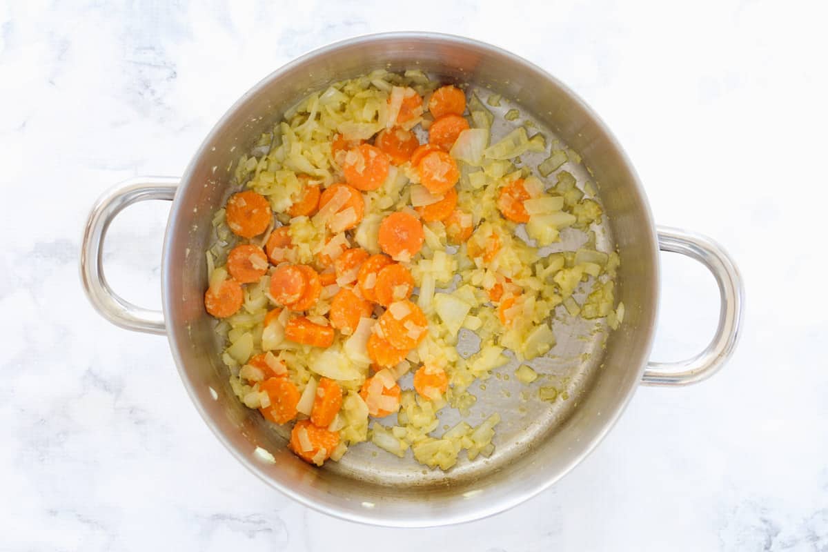 A stainless steel casserole pan with diced onions, minced garlic and sliced carrots sautéed in butter.