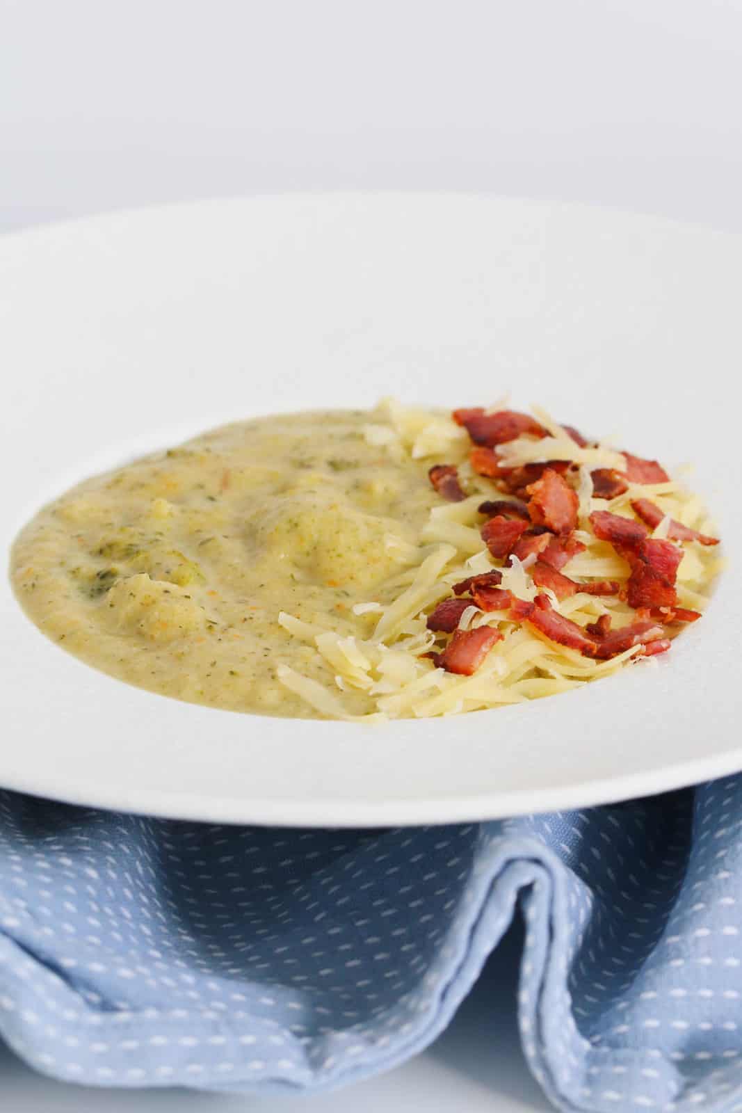 A white soup plate on a blue and white tea towel with a thick, chunky broccoli and cauliflower soup topped with bacon and cheese.