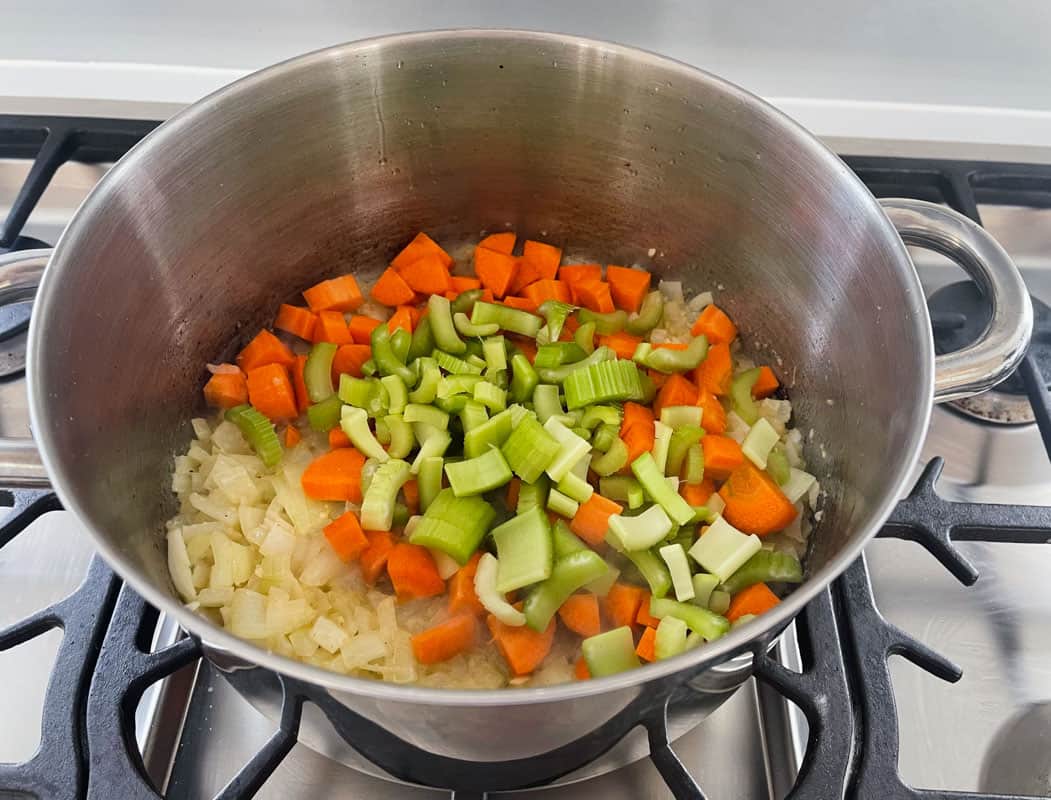 Chopped carrots and celery added to a stainless steel pot with translucent sauteed diced onions