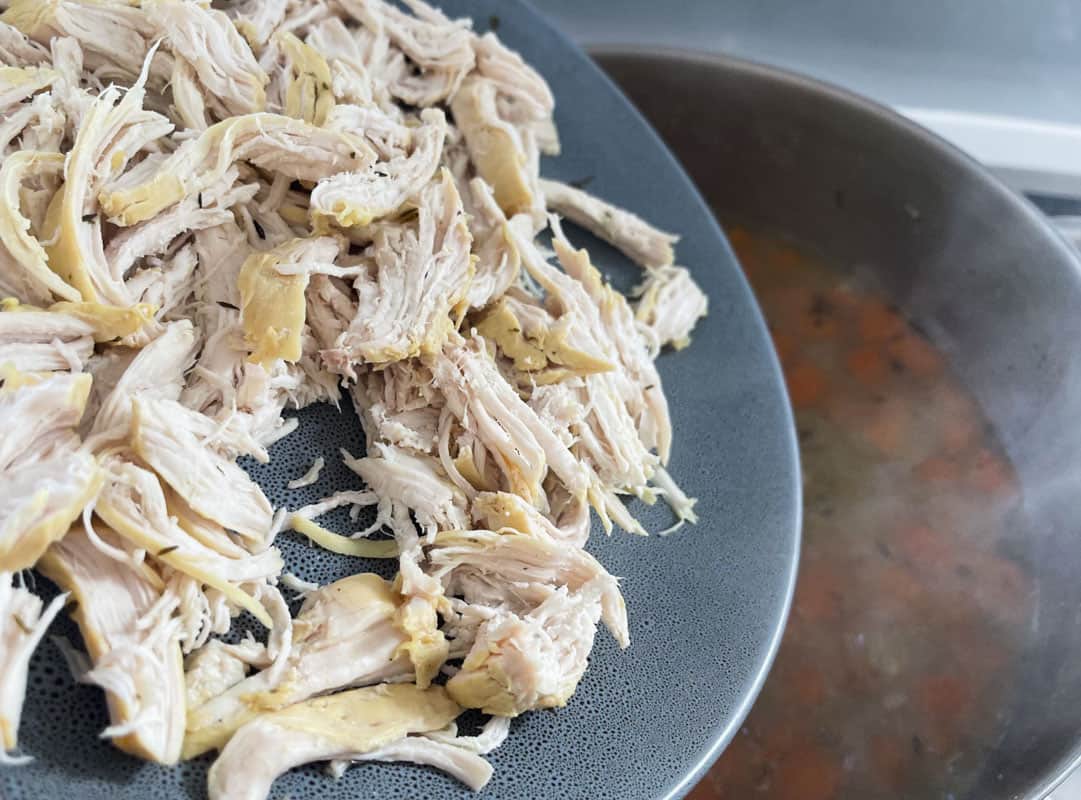 Coarsely shredded chicken on a grey plate being added back into a blurred pot of stock and vegetables below
