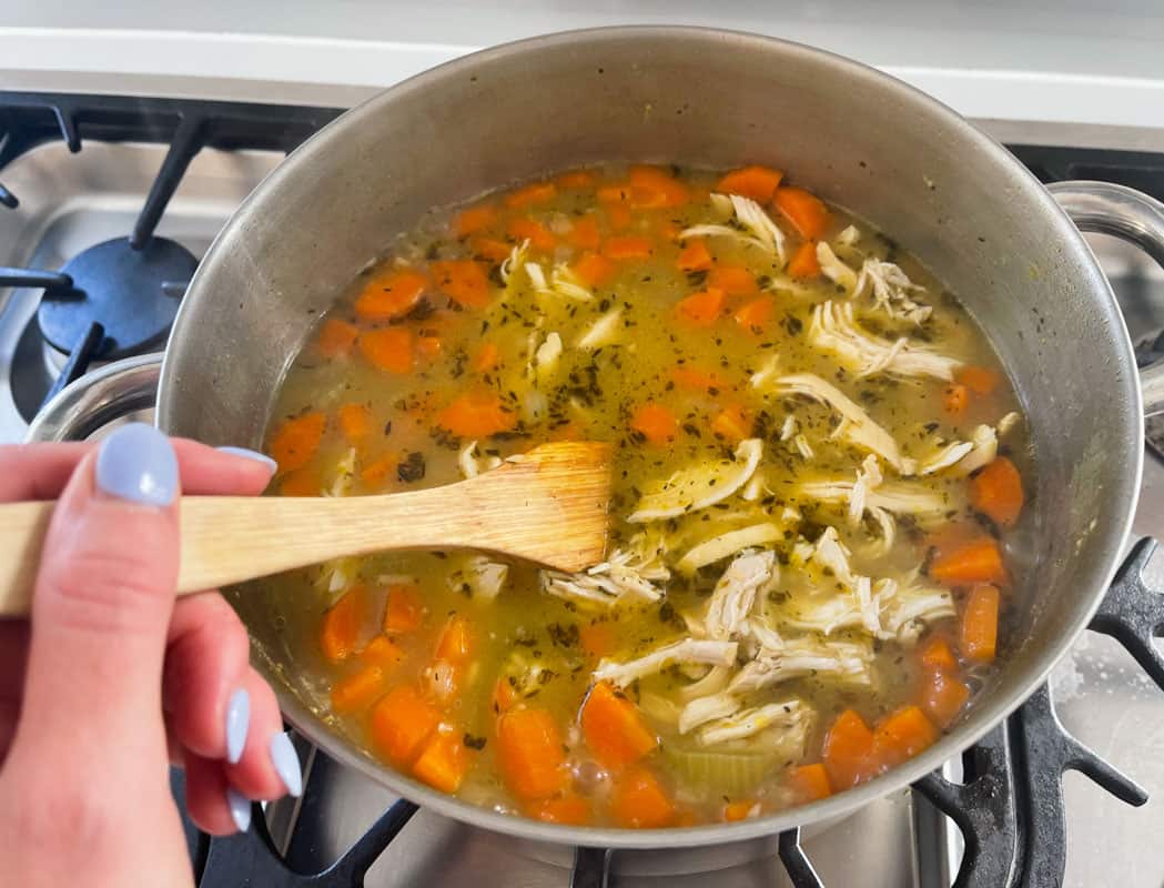 A wooden spoon being used to stir a soup with shredded chicken and vegetables