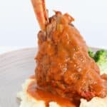 A lamb shank covered in a rich tomato gravy arranged on a bed of mash with broccoli in the background.