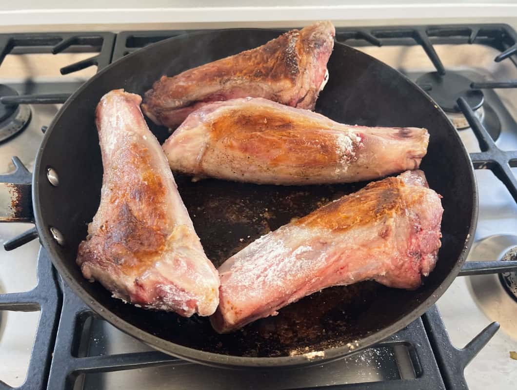 Four lamb shanks being seared in a non stick pan on a stovetop.