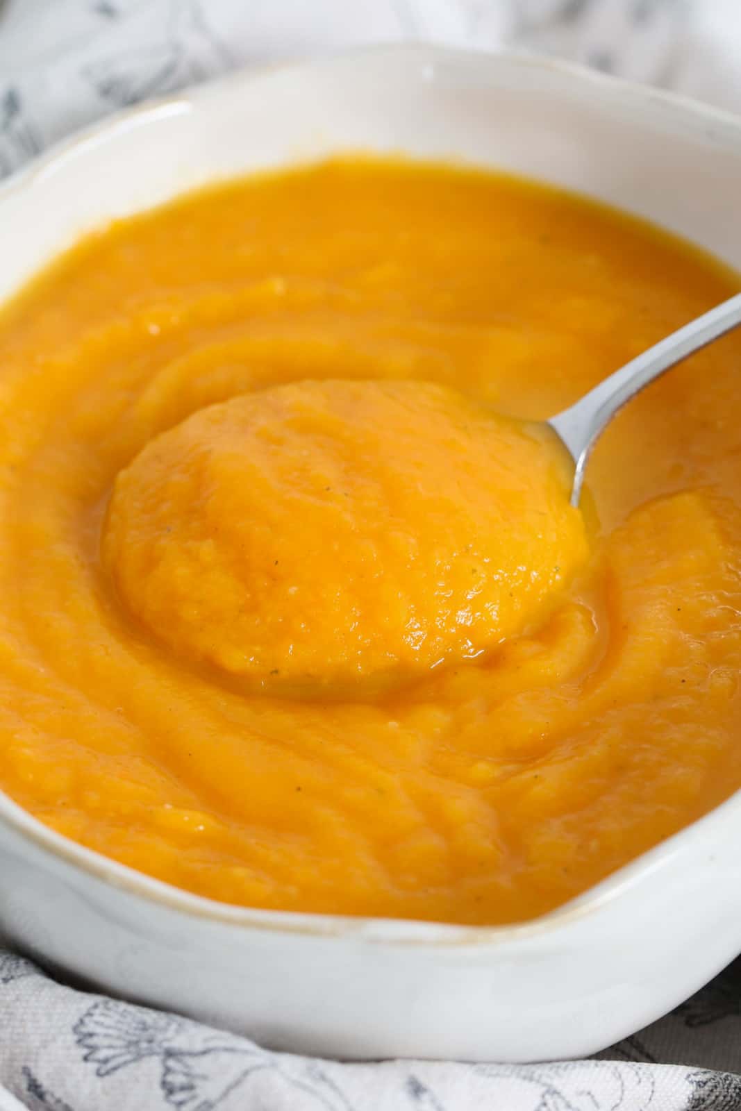 A close up view of a white bowl with a soup spoon lifting a spoonful of thick orange coloured soup.