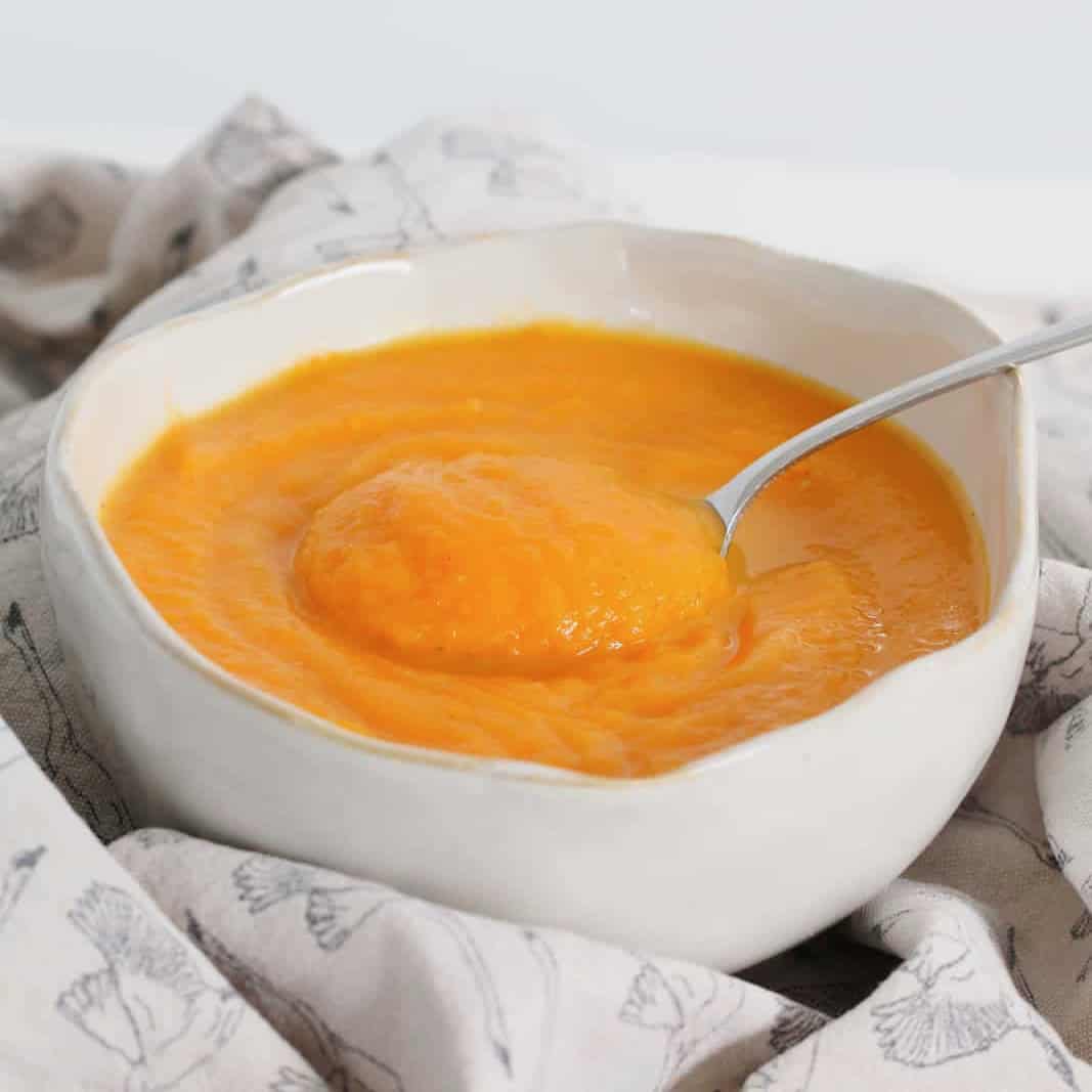 Carrot, Pumpkin & Sweet Potato Soup in a white bowl surrounded by a patterned tea towel