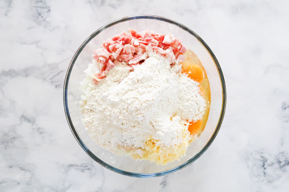 A glass bowl on a marble counter with flour, bacon, cheese and eggs waiting to be combined.