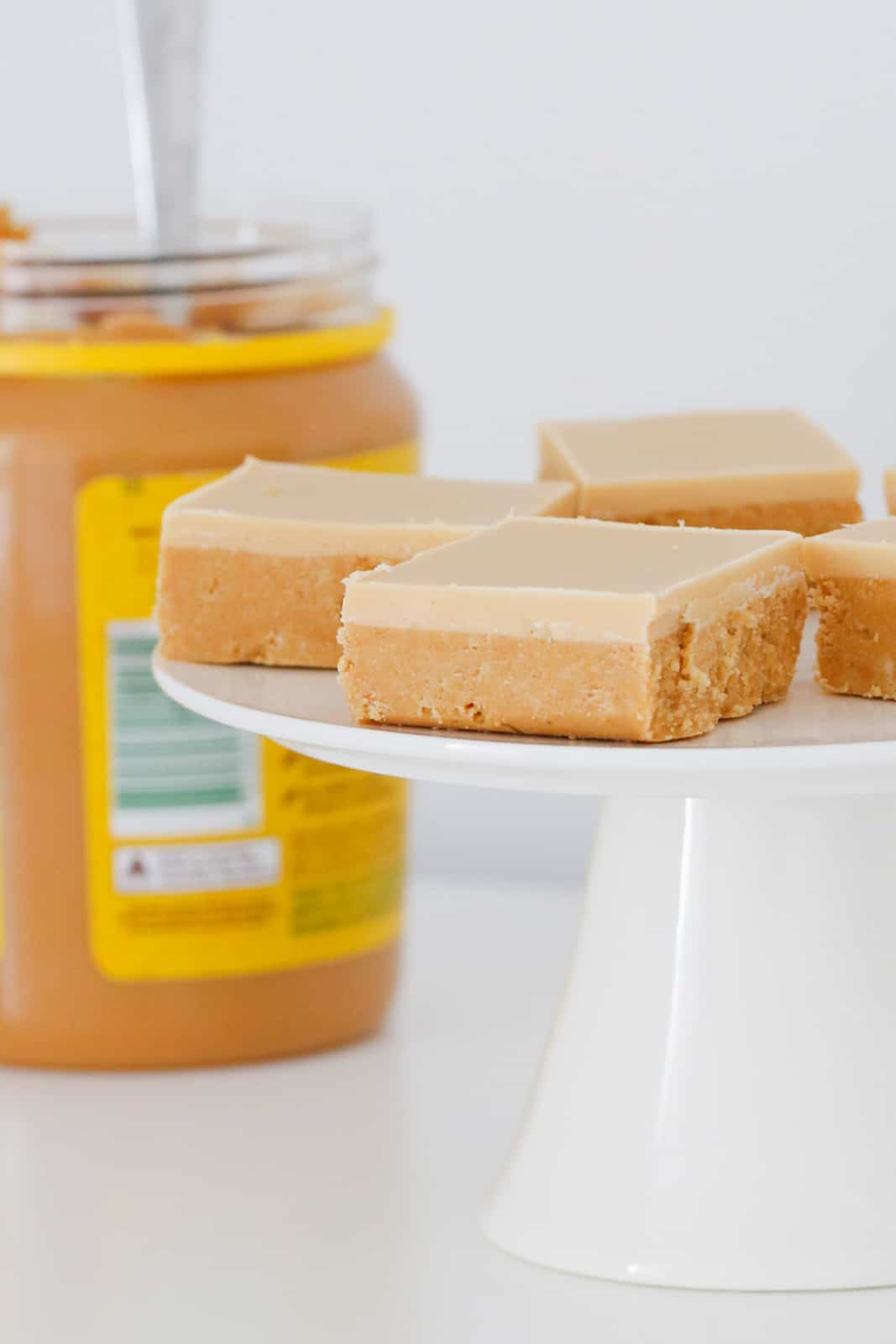 A jar of peanut butter behind a cake stand topped with squares of slice topped with a white chocolate and peanut butter icing.