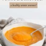 A white bowl of thick pumpkin, sweet potato and carot soup, placed on a tea towel.
