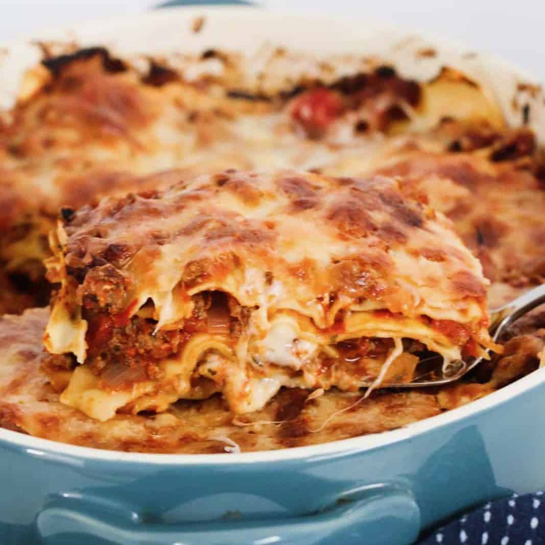 a baking dish of lasagne, with a tea towel wrapped around it, with a serving being lifted out