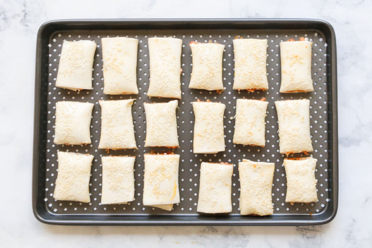 A crisping baking tray on a marble counter with raw sausage rolls arranged on it