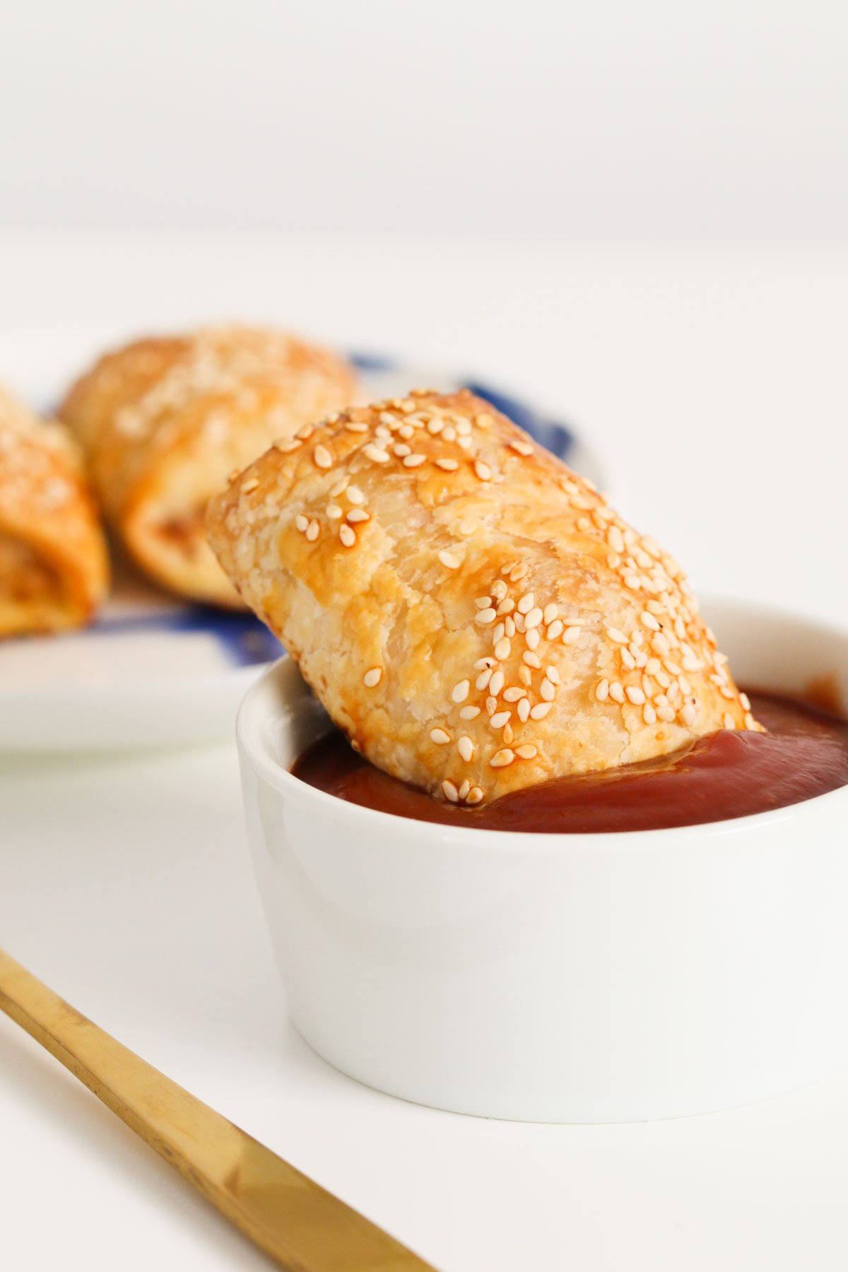 A puff pastry roll covered in sesame seeds with one end dipped in a small bowl of tomato sauce.