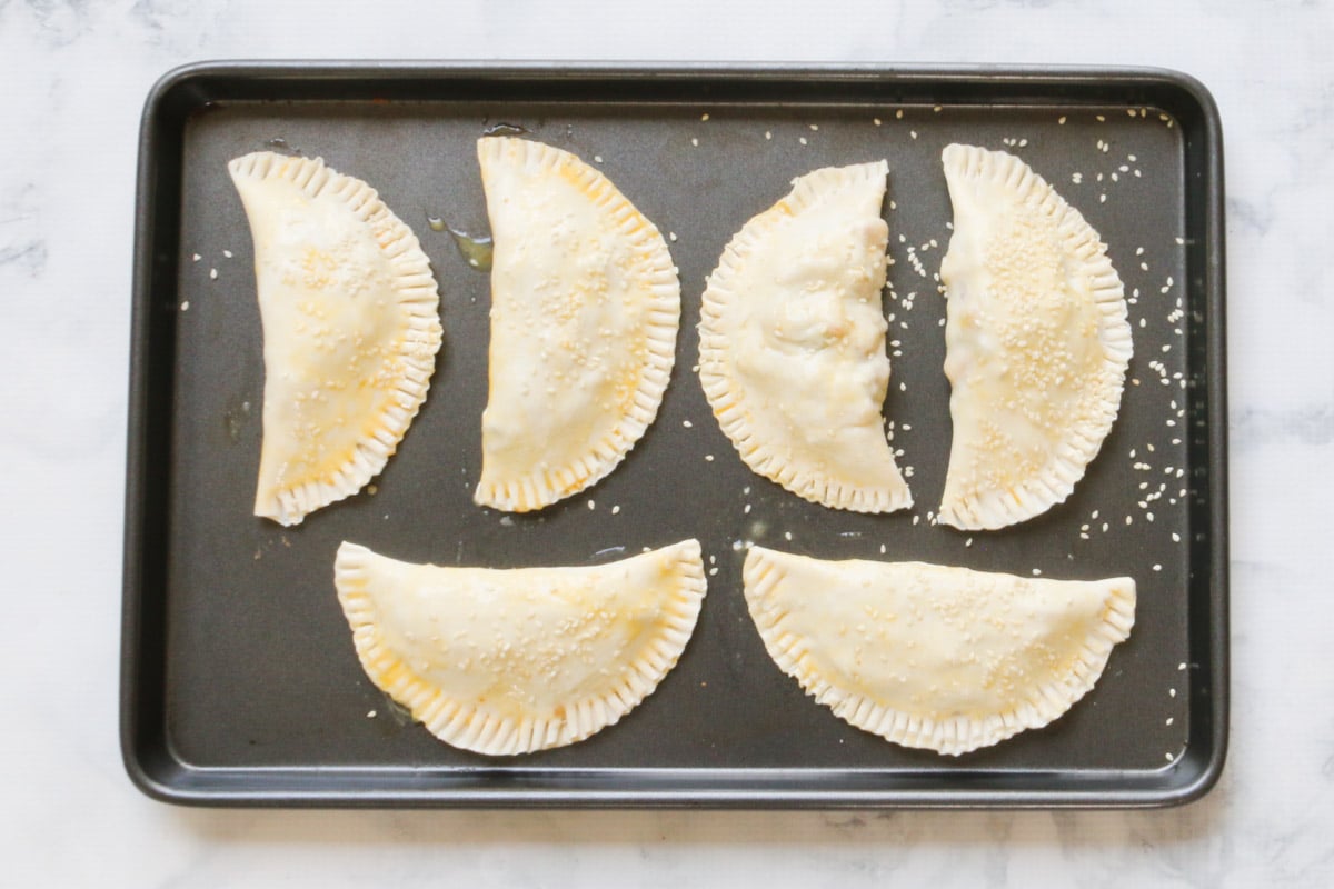 A baking tray with 6 unbaked pasties on it, each brushed with egg wash, and sprinkled with sesame seeds.