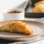 a single pastie on an earthy-coloured plate with a ramekin of tomato sauce and more pasties in the blurred background