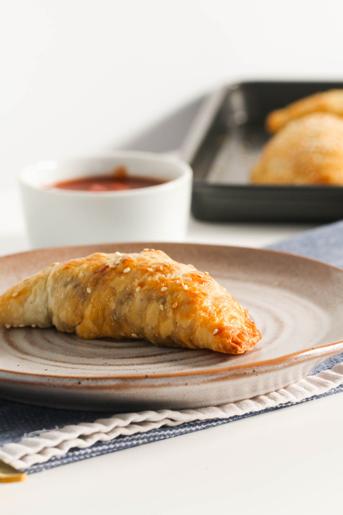 A single baked pastie on an earthenware plate with a ramekin of tomato sauce and more pasties in the blurred background